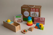 Montessori toy box set with a brown cardboard box, wooden baby tray, colorful stacking rings, blocks, and cards on a neutral beige background for a baby aged 9-10 months