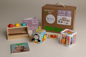 Montessori sensory play box set by Sensory Stories, including a brown cardboard storage box, wooden shelf with colorful balls, penguin activity cube, colorful wire cage toy, and cards, arranged on a beige background for a baby aged 11-12 months