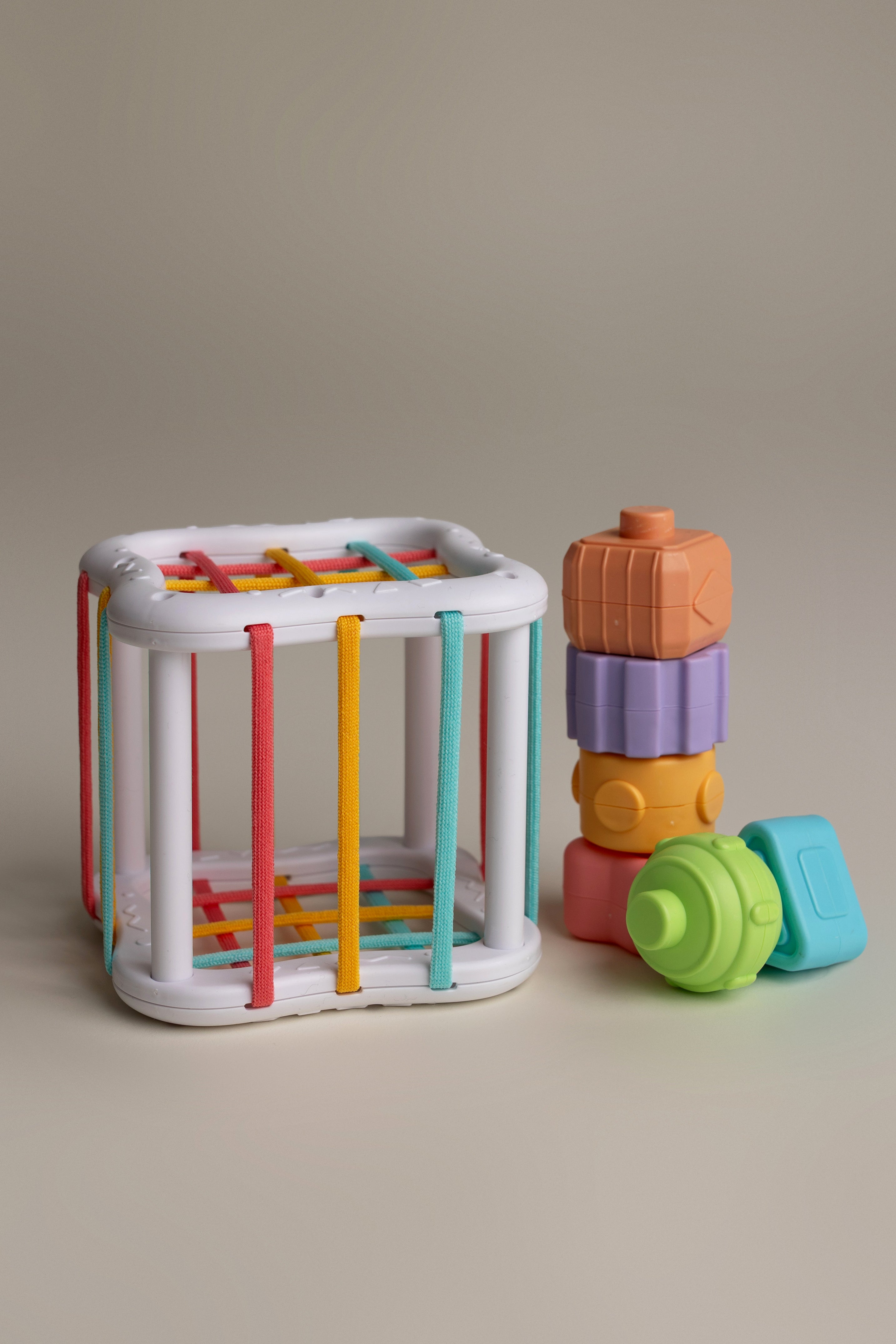 Colorful baby toys including a white activity cube with colorful strings and a stack of stacked blocks on a beige background.