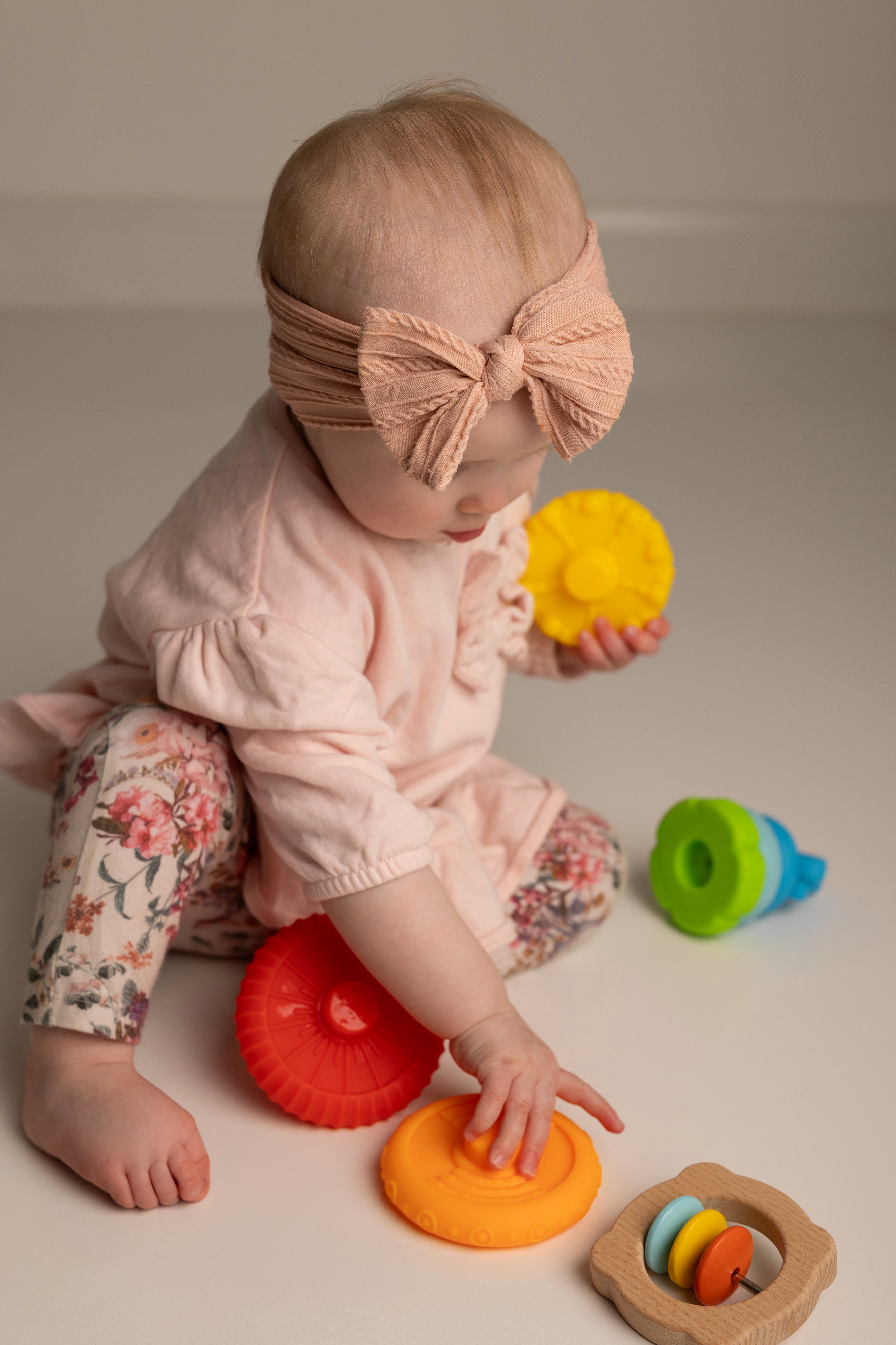 A baby girl wearing a pink ruffled top, floral pants, and a matching pink bow headband sits on a light beige floor playing with colorful wooden toy rings.
