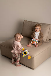 Two babies playing with a toy on a beige sofa in a minimalistic room.