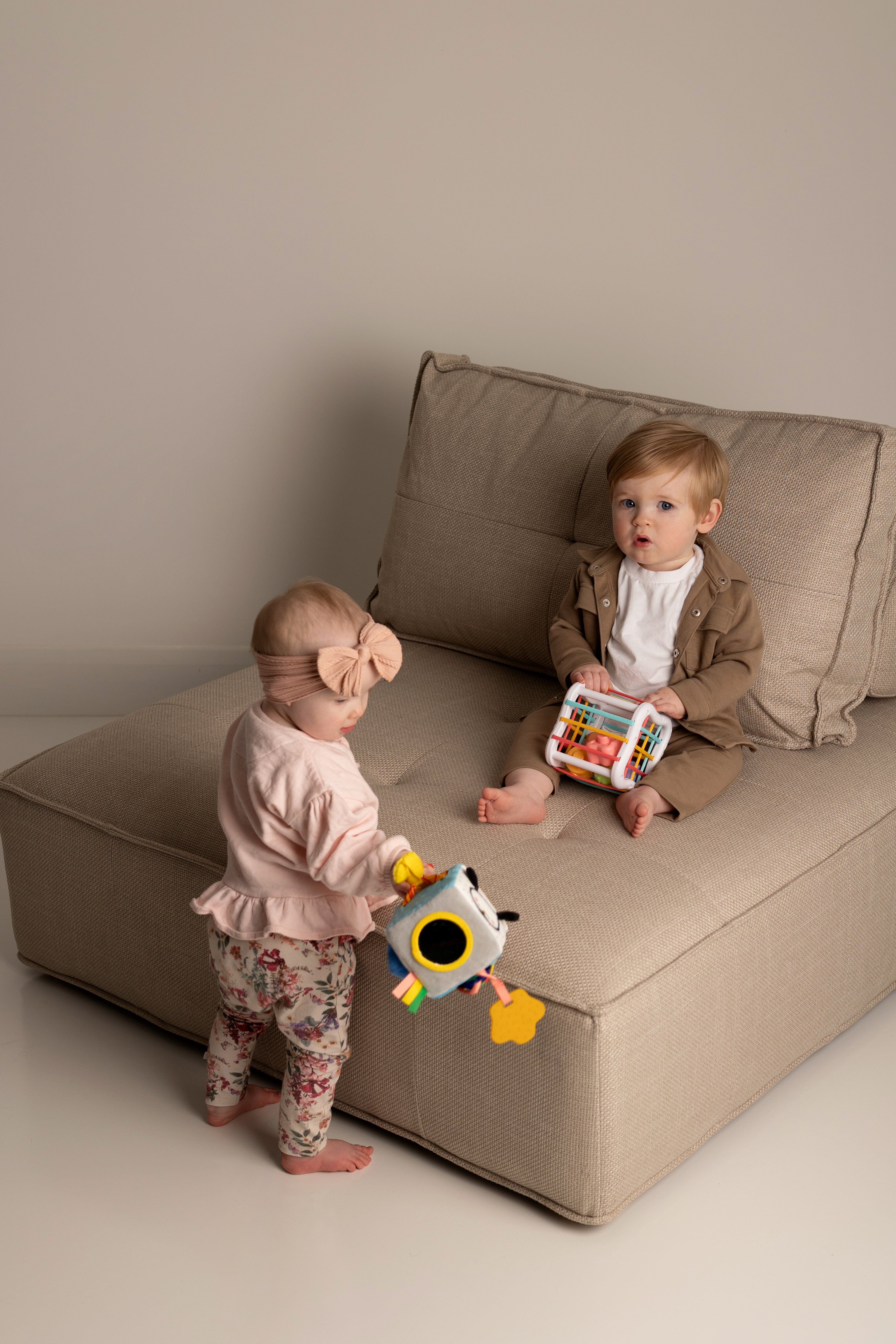 Two babies playing with a toy on a beige sofa in a minimalistic room.